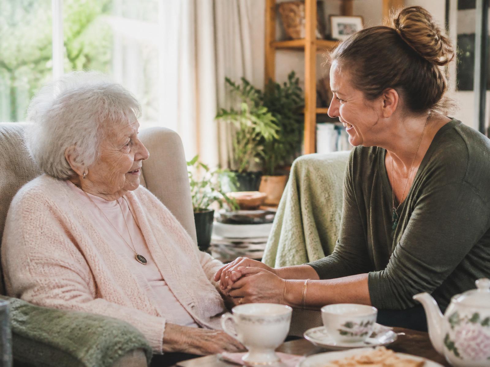 A caregiver sharing tea and warm conversation with an elderly woman in her sunlit living room